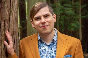 Headshot of Basil, a white nonbinary person with an undercut, wearing a mustard blazer and patterned blue shirt.