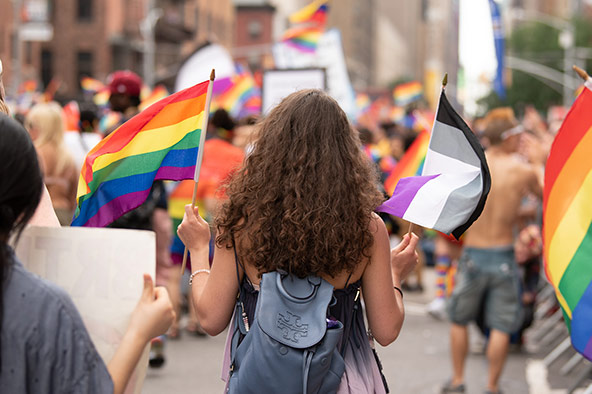 A person with light skin and long, curly brown hair holds a pride flag and an ace flag as they march in a parade.