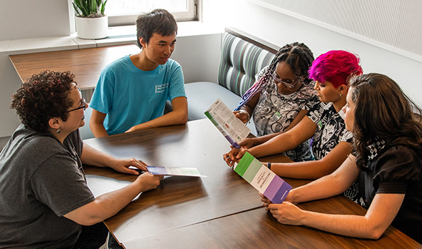 Five people with various skin colors are sitting around a table reading green and purple brochures.