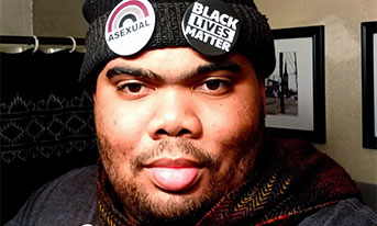 A Black man with facial hair wearing a beanie with "Black Lives Matter" and "Asexual" pins holds up two fingers.