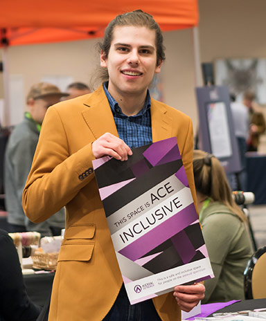 A white person in a mustard jacket holds a purple poster that says "This Space is Ace Inclusive"