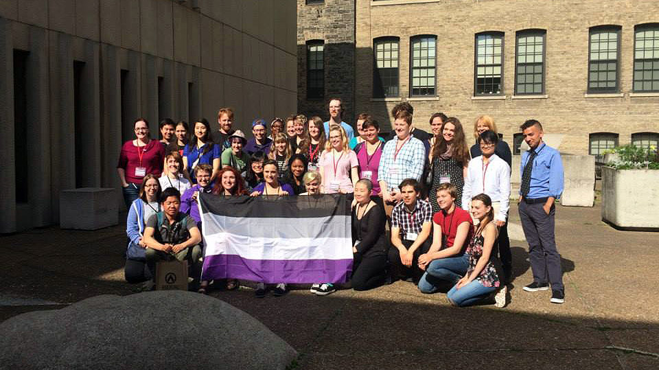 Three dozen people pose as a group behind an ace flag. They are in a courtyard surrounded by buildings.
