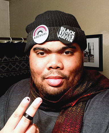 A Black man with facial hair wearing a beanie with "Black Lives Matter" and "Asexual" pins holds up two fingers.