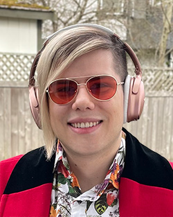 Headshot of Basil, a white nonbinary person with a blonde undercut, wearing red glasses and pink headphones.