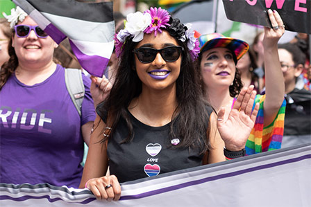 Three people march in a pride parade behind a banner. The one in front has brown skin and is waving.